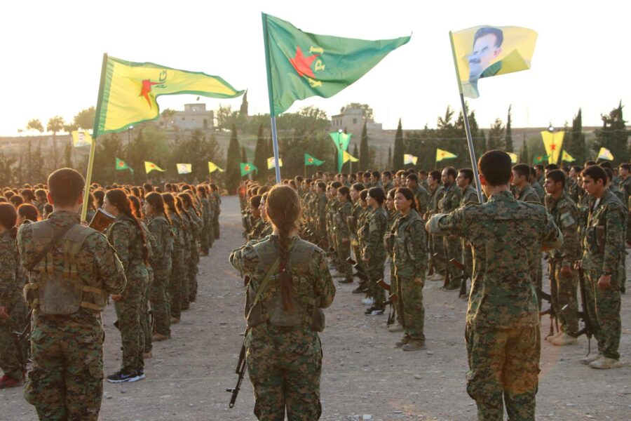 Man sieht mehrere Reihen kurdische Kämpfer*innen mit Fahnen der YPG, YPJ und von Öcalan.