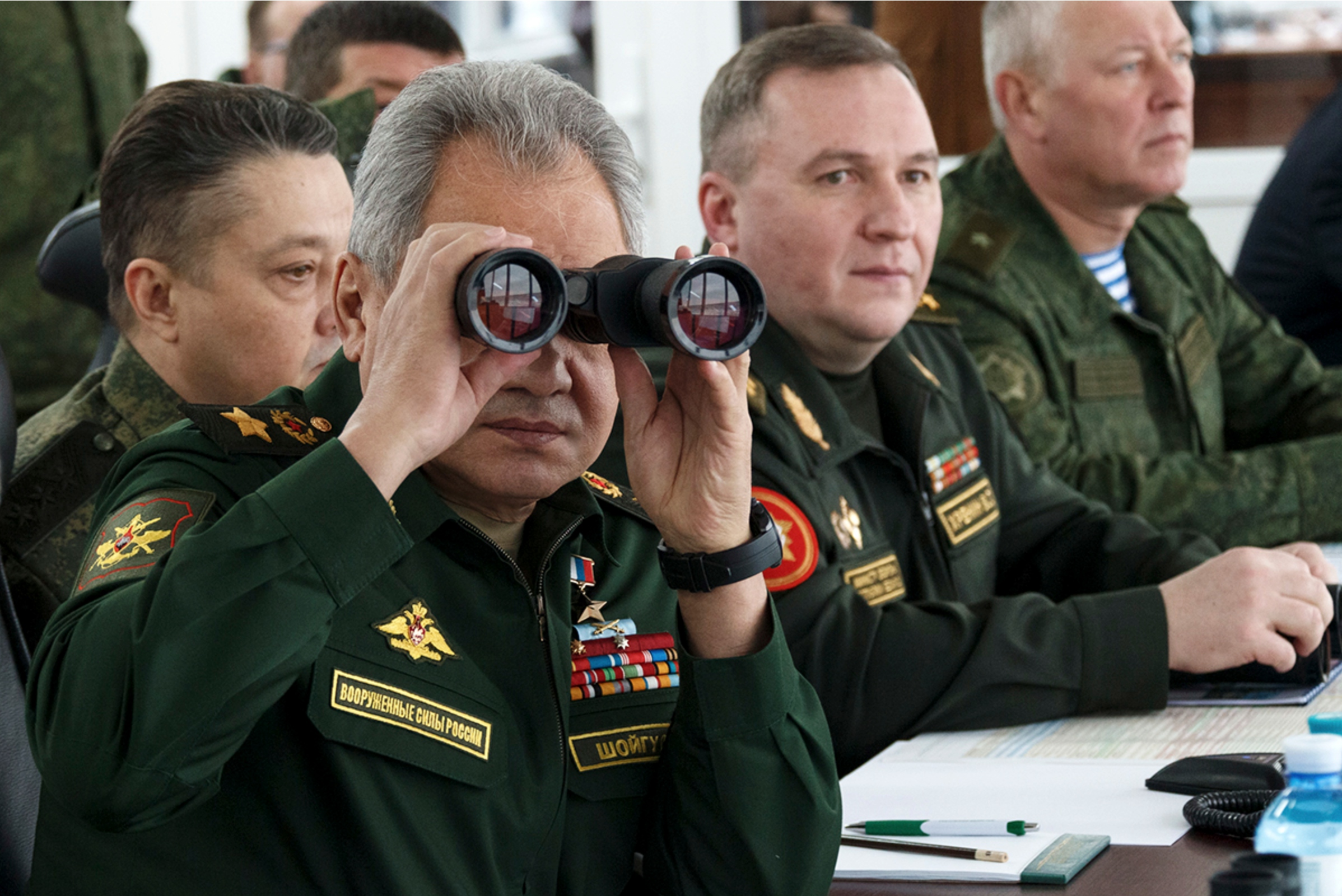 Ein grauhaariger Mann in Militäruniform sitzt an einem Tisch und sieht durch ein Fernglas, hinter ihm weitere Männer in Militäruniform