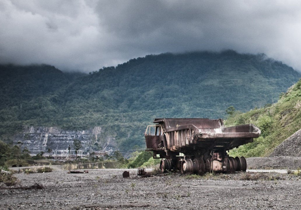 Bild einer Landschaft. In der Ferne erheben sich Hügel, die in tiefsitzenden Wolken verschwinden. Vorne die Überbleibsel einer alten Mine, viel Schotter, in der Mine die verrosteten Überbleibsel eines LKW