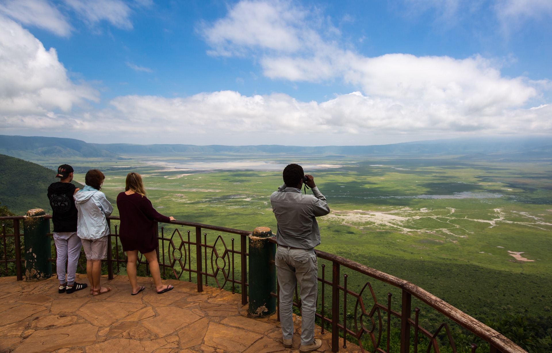 Panorama-Bild einer weiten Graslandschaft mit einem Fluss, der durch die Mitte fließt. An den Rändern sind Gebirge. Vorne im Bild stehen Vier Tourist*innen auf einem Deck und schauen in die Ferne.