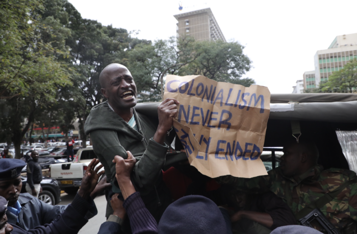 Man sieht eine Person, die von Polizisten verhaftet wird, in der Hand hält die Person ein Schild, auf dem steht: colonialism never ended.