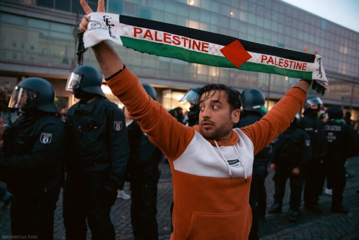 Ein Mann hält einen Schal in die Luft, auf dem zweimal der Schriftzug "Palestine" zu lesen ist. Er Blickt hinter sich. Dort stehen, mit dem Rücken zu ihm, eine Reihe behelmter Politzist*innen