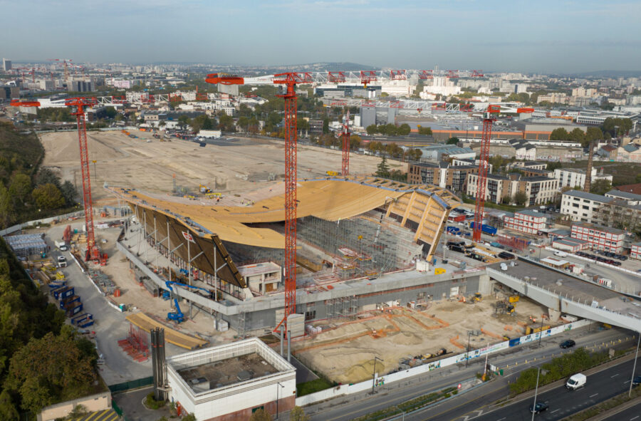 Luftbildaufnahme einer Baustelle für ein Stadion, mit roten Kränen, einem Fundament aus Beton und einer Holzverkleidung obendrauf.