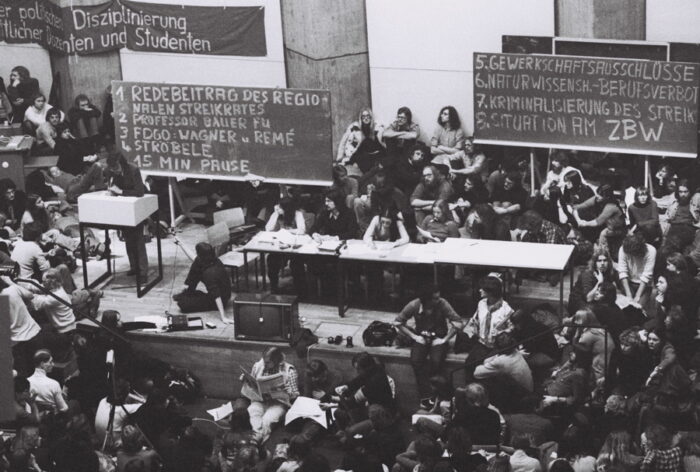 Auf dem schwarz-weiß Foto ist eine Szene aus den 1970er Jahren zu sehen. Es scheinen überwiegend Studierende zu sein, die in einem vollen Saal auf zwei Tafel blicken. Als ein Tagesordnungspunkt ist dort unter anderem "Gewerkschaftliche Unvereinbarkeitsbeschlüsse" aufgelistet