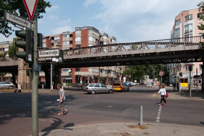 Zwei Menschen überqueren eine Straße, im Hintergrund eine Hochbahn. Vorn sieht man zwei Straßenschilder, auf einem steht "Bülowstraße", auf dem anderen "Frobenstraße"