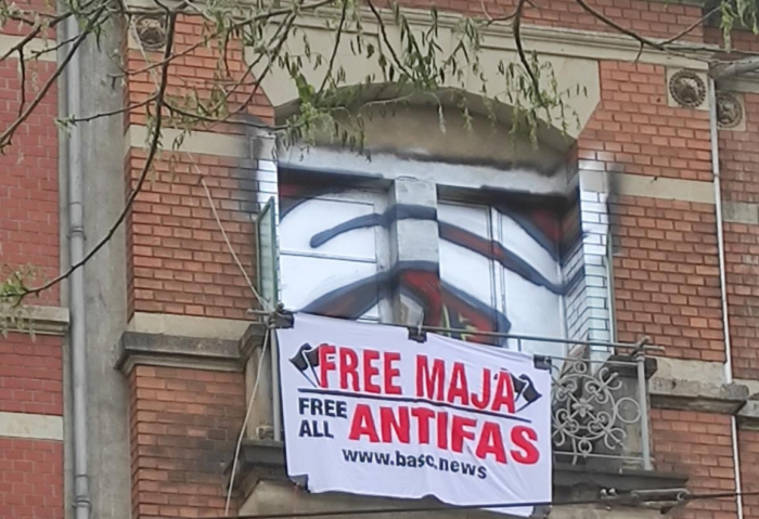Ein Banner häng über dem verschnörkelten Balkongeländer eines alten Hauses. Die Fensterläden im Hintergrund sind mit Graffiti besprüht. Auf dem Banner steht: Free Maja. Free all Antifas. www.basc.news.