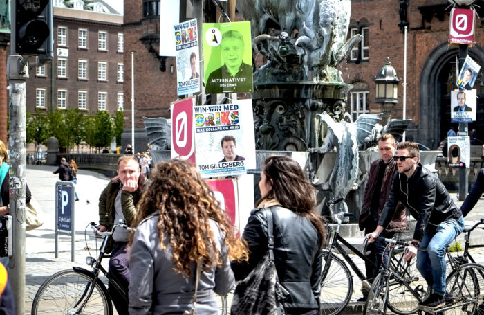 Straßenszene mit Radfahrer*innen und Fußgänger*innen in einer nordischen Altstadt, überall hängen Wahlplakate