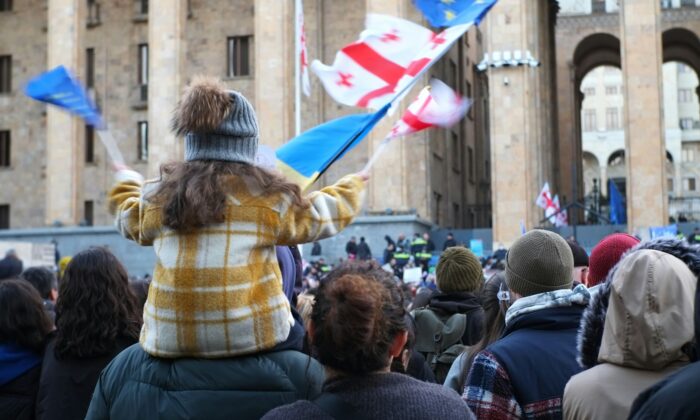 Menschenmenge (von hinten fotografiert) vor einem Gebäude mit Säulen. Im Vordergrund ist ein Mädchen mit Mütze zu sehen, das auf den Schultern einer anderen Person sitzt, man sieht Fahnen von Georgien, der Ukraine und der EU.