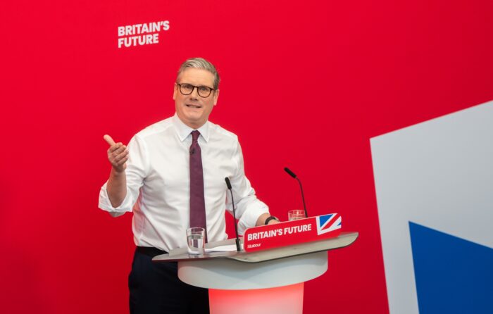 Keir Starmer steht an einem Rednerpult und schaut ins Bild, auf dem Pult und an der roten Wand dahinter steht "Britain's Future"
