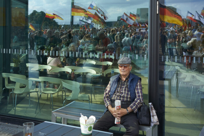 Ein älterer Mann in Hemd und Weste sitzt allein vor einem Café, in der Scheibe des Cafés spiegelt sich eine Demonstration mit Deutschland-, Berlin- und Russland-Fahnen, auch ein Reichsadler ist zu sehen.