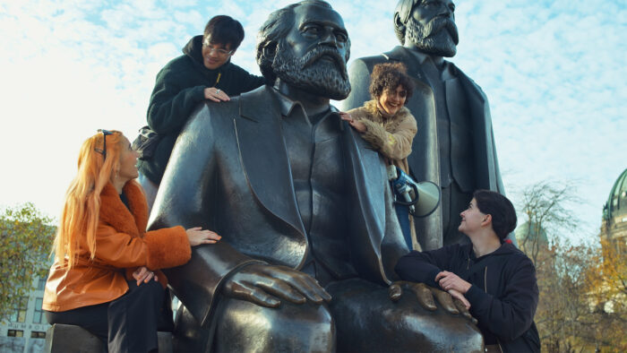 Man sieht ein Denkmal von Karl Marx und Friedrich Engels, auf dem vier Personen lachend herumklettern.