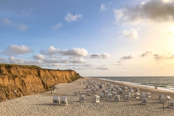 Das Bild zeigt das rote Kliff auf Sylt. Daneben ist ein weißer Sandstrand mit weißen, menschenleeren Strandkörben. Am blauen Himmel sind einige Wolken zu sehen. Zwei Möwen kreisen über den Strandkörben.