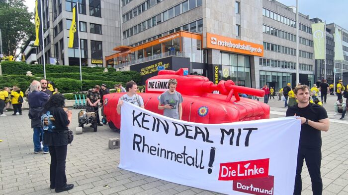 Vor dem BVB-Fanshop in Dortmund stehen Mitglieder der Partei Die Linke. Sie haben einen großen roten, aufblasbaren Panzer dabei. Drei junge Männer halten ein Transparent auf dem steht: Kein Deal mit Rheinmetall. Im Hintergrund sind einige BVB-Fans zu sehen.