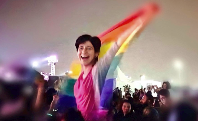 Eine Person hält eine Regenbogenflagge über einer Menschenmenge und lacht ausgelassen.