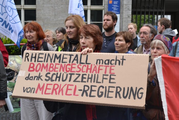 Ein Gruppe von Demonstrierenden hält ein Schild mit der Aufschrift "Rheinmetall macht Bombengeschäfte mit Schützenhilfe der Merkel-Regierung.