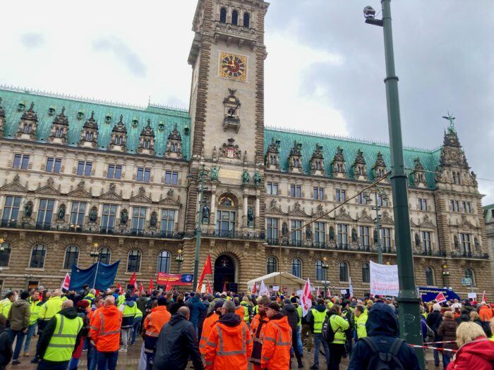 Etwa 200 Menschen, viele in gelben ver.die Warnwesten oder organgenen Arbeitswesten, stehen vor dem Hamburger Rathaus und demonstrieren. Eine halten Protestplakate. Es ist bewölkt.