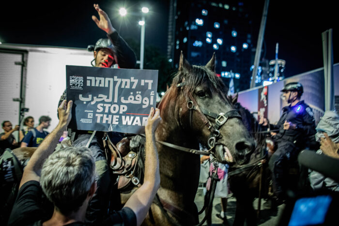 Ein Polizist auf einem Pferdam späten Abend am Rande einer Demonstration, er macht eine Handbewegung in Richtung einer Demonstrantin. Die ältere Frau im Vordergrund, die man von hinten sieht, hält ein Schild hoch, auf dem "Stop the War" steht.