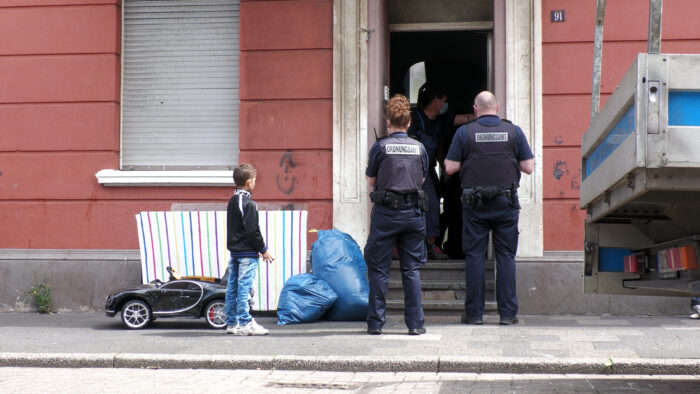 Zwei Mitglieder der Taskforce Problemimmobilie, von hinten aufgenommen, stehen an einem Hauseinang vor einer offenen Tür. Eine Person im inneren des Hauses spricht mit ihnen. Neben den Beamt*innen steht ein kleineres Kind und beobachtet die Szenerie.