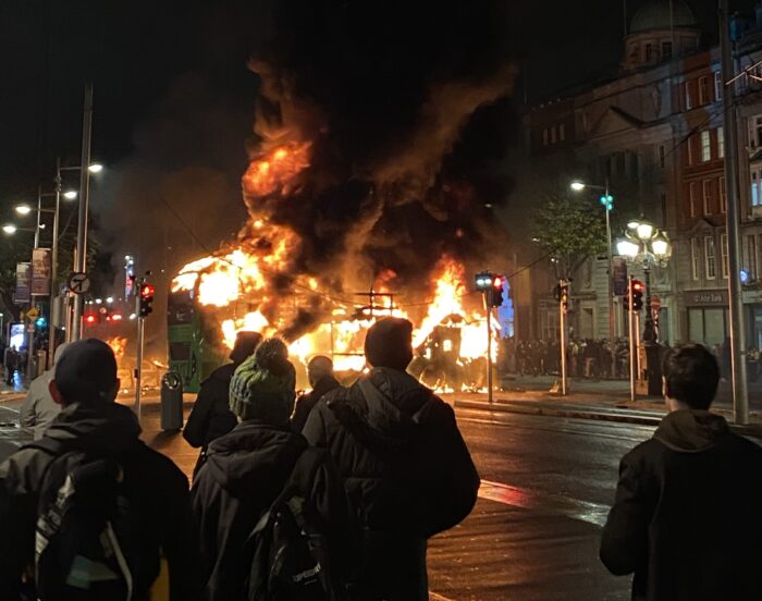 Eine Gruppe von hinten fotografiert, die dabei zu schaut, wie ein Bus brennt.