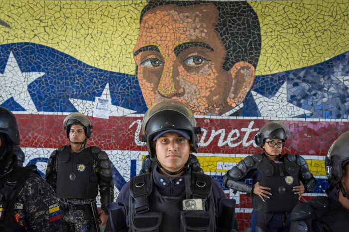 Das Bild zeigt drei Mitglieder der Riot Police in Caracas, einer von ihnen sieht direkt in die Kamera. Hinter ihnen ist ein Wandgemälde zu erkennen, vermutlich von Rafael Urdaneta. Der General kämpfte an der Seite des Nationalhelden Simon Bolivar.