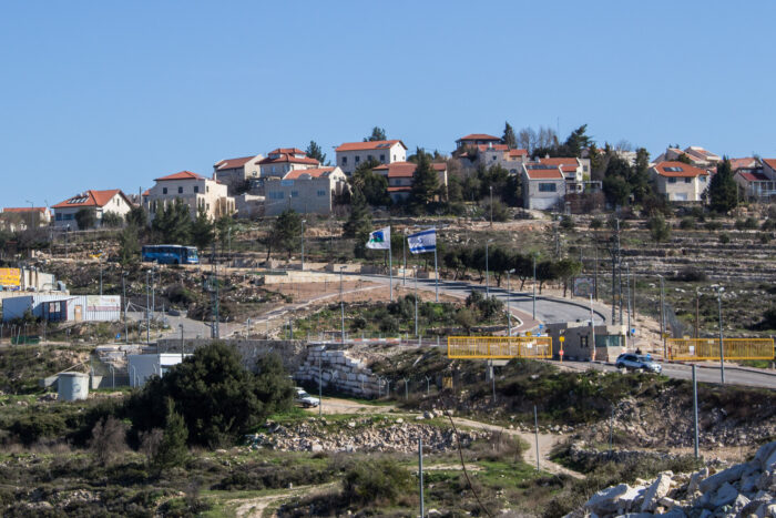 Blick auf eine Reihe Häusern an der Spitze eines Hügels, umgeben von Zäunen. In der Mitte weht eine israelische Flagge