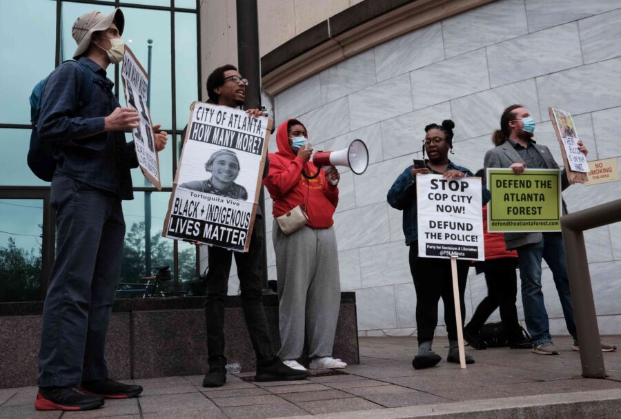 Demonstrant*innen mit Schildern stehen vor einem Gebäude mit Glasfront. Auf den Schildern steht: "Stop Cop City", "Defund the police" und "City of Atlante, how many more? Black and Indigenous Lives Matter".