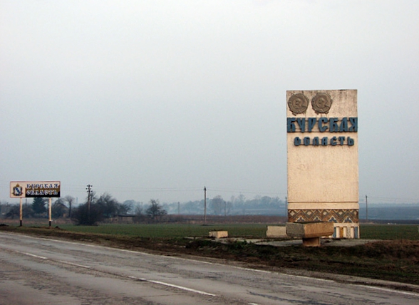 Landstraße bei regnerischem Wetter, am Straßenrand steht eine Stele mit der Aufschrift Курская область (Kursk Oblast).