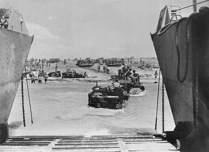 Ein Foto aus einem Landungsboot heraus, auf einen Strand, dort Panzer, Soldaten und Militärfahrzeuge