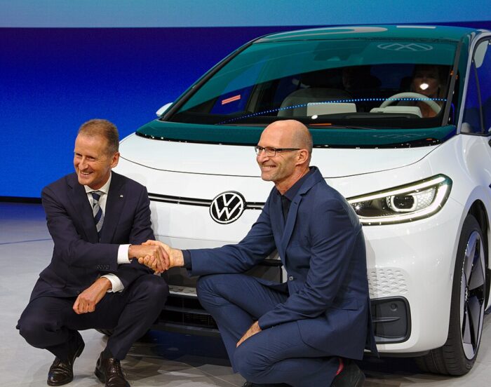 Der ehemalige VW Manager Herbert Diess und der Automobildesigner Klaus Zyciora knien auf einer Bühne vor einem weißen neuen VW. Sie grunsen in die Kamera und schütteln sich die Hände. Das Bild entstand 2019 auf der Internationalen Automobilausstellung IAA