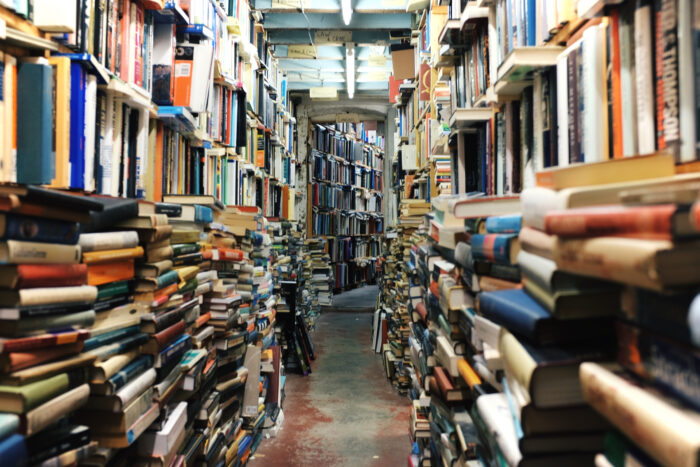 Blick durch einen Gang voller Buchstapel und Bücherregale in einen anderen Raum, in dem weitere Bücherregale stehen