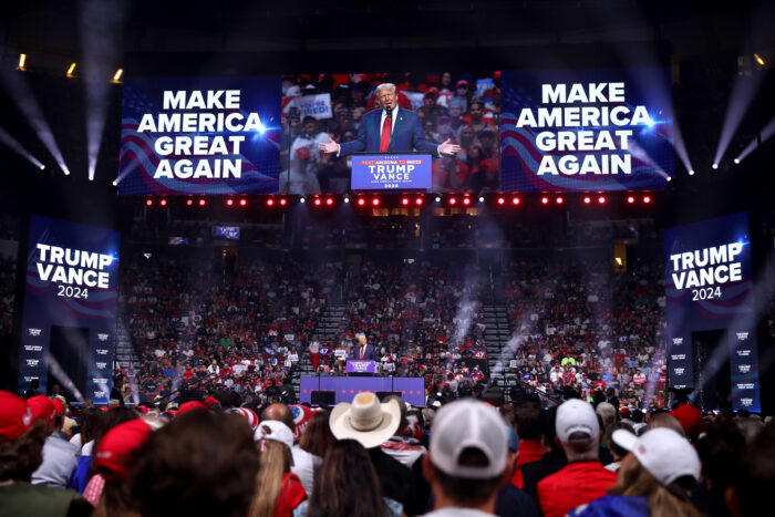 Donald Trump steht bei einer Wahlkampfveranstaltung in einem Ring auf einer Bühne, über der Bühne hängt ein großer Bildschirm mit Trump und dem Slogan Make America Great Again, vor der Bühne sind viele Menschen mit Caps und Cowboyhüten zu sehen