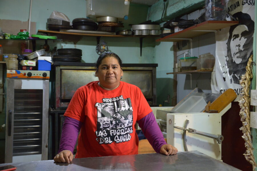 Die Aktivistin Mónica Troncoso steht hinter dem Tresen einer genossenschaftlichen Pizzeria in Buenos Aires. Sie trägt ein rotes T-Shirt mit Aufrduck und Zopf und guckt in die Kamera. Hinter ihr sind Regale zu sehen, mit Töpfen und Pfannen