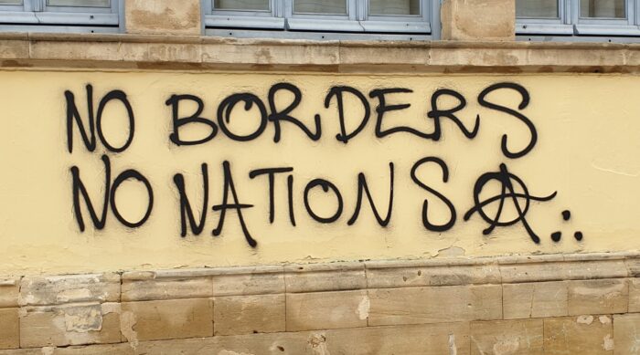 Graffiti an einer beigen Wand: No Borders No Nations, danach ein Anarchie A