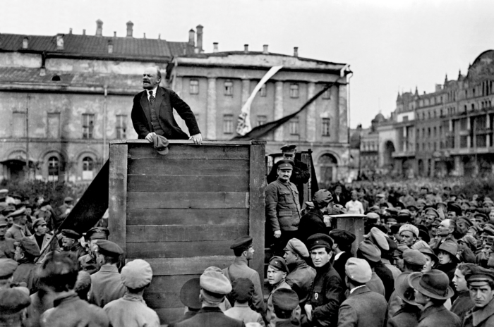 Man sieht ein Holzpodest, das umgeben ist von vielen Menschen. Auf dem Podest steht Lenin und hält offenbar eine Rede.