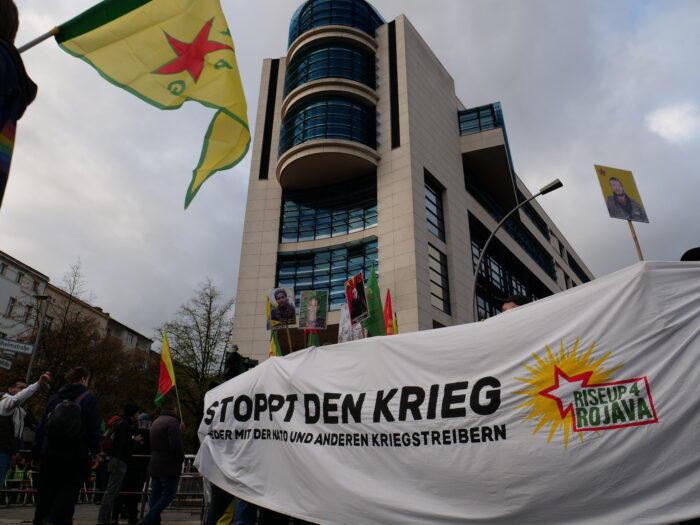 Banner auf einer Demonstration mit der Aufschrift "Stoppt den krieg - Nieder mit der Nato und anderen Kriegstreibern" und dem Logo der Initiative RiseUp4Rojava. Oben links im Bild ist eine Fahne der Volksverteidigungseinheiten YPG, weiter hinten Schilder von in Rojava gefallenen Kämpfer*innen.