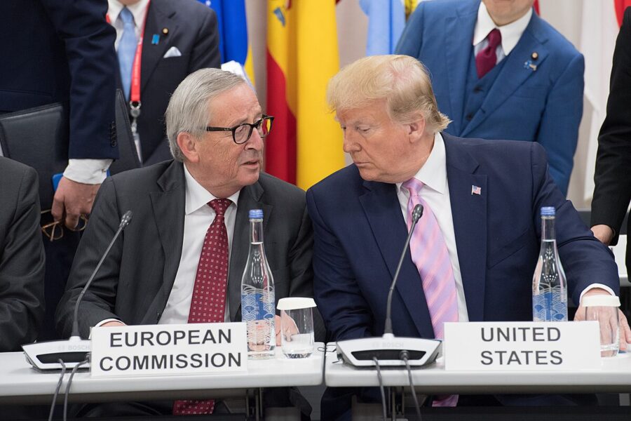 Zwei Männer mit Krawatte, Jean-Claude Juncker und Donald Trump, sitzen nebeneinander an einem Verhandlungstisch, vor ihnen Schilder mit der European Commission und United States, hinter ihnen Männern mit Krawatte.