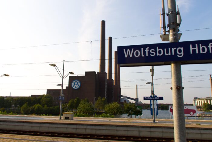 Im Vordergrund ein Bahnhofsschild mit der Aufschrift "Wolfsburg Hbf", im Hintergrund das VW-Werk.