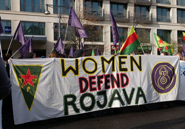 Banner auf einer Demonstration mit der Aufschrift »Women Defend Rojava«, links daneben das Emblem der Frauenverteidigungseinheiten YPJ und rechts davon das Logo der feministischen Vernetzung Women Defend Rojava