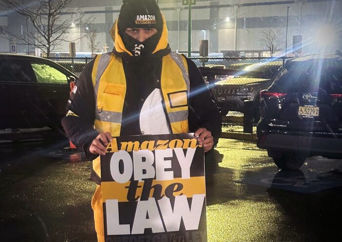 Ein dick angezogener Mann mit Warnwester und Teamsters-Mütze steht bei Dunkelheit auf einem beleuchteten Parkplatz. Er schaut in die Kamera und hält ein Schild mit der Aufschrift "Amazon, obey the law".