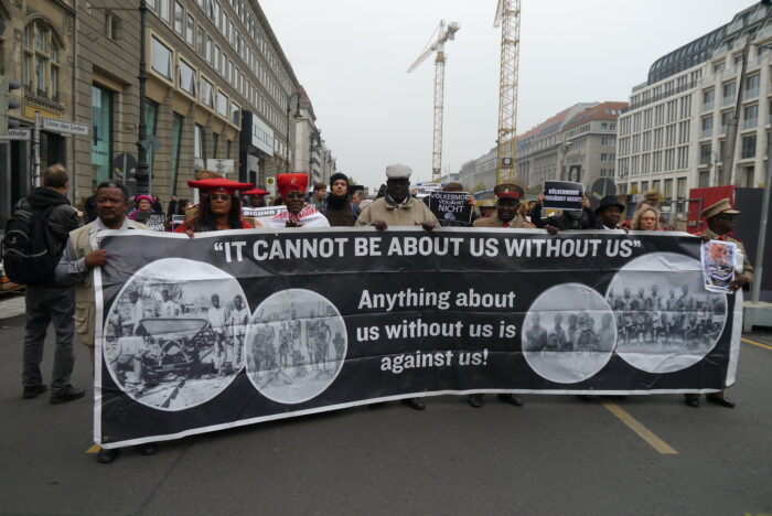 Foto eines Demonstrationszuges. In der erste Reihe ein Plakat mit dem Text "It cannot be abot us without us. Anything about us without us is against us"