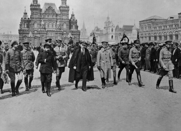 Ein Gruppe von Männnern in Uniformen und Anzügen, schreitet eine Reihe von Soldaten ab, dahinter ein Gebäude mit vielen Türmen