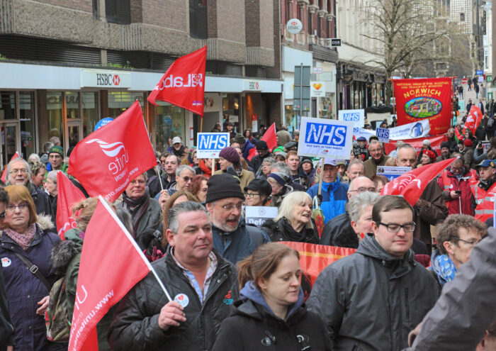 Das Foto zeigt einen Protestzug 2017 im britischen Norwich. Einige Menschen tragen rote Gewerkschaftsfahnen, andere Plakate mit der Aufschrift NHS, not safe in Tory hands