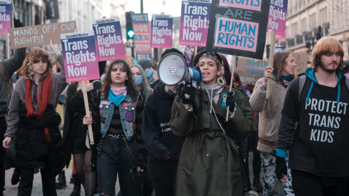 Man sieht viele Personen bei einem Protest, sie halten Schilder hoch auf denen steht Trans Right now. Eine Person hat ein Megaphon.