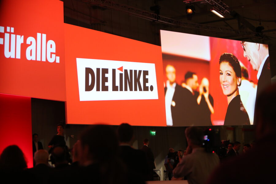 Man sieht eine Parteitagsbühne, an die Wand ist "Die Linke" projiziert und daneben sieht man Wagenknechts Gesicht in Großaufnahme.