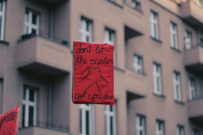 Schild vor Berliner Hauswand, auf dem zwei gemalte Hände ineinandergreifen, dazu steht in Handschrift: Don't let the system get you down