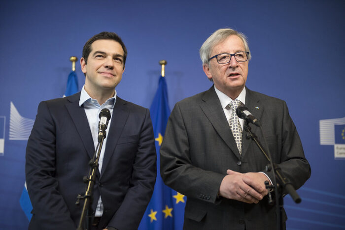 Zwei Männer (links Alexis Tsipras, rechts Jean-Claude Juncker im Anzug stehen neben einander, der rechte spricht in ein Mikrofon, dahinter steht eine EU-Fahne.