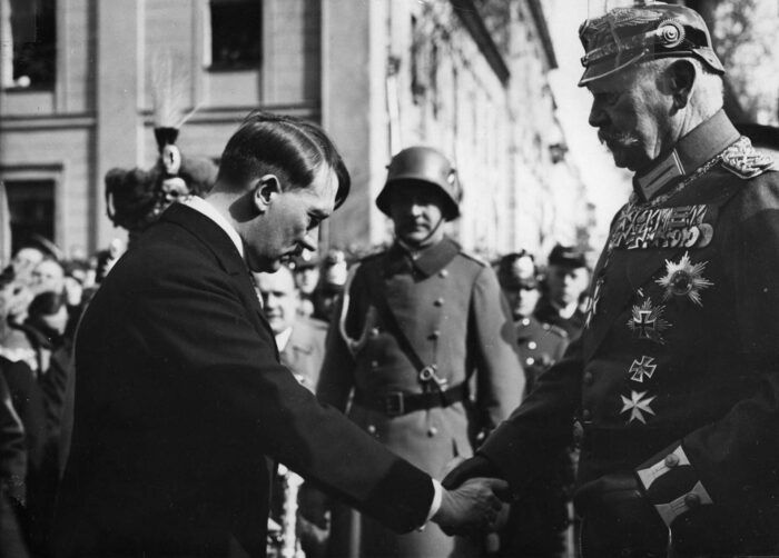 Ein Mann in im Anzug verbeugt sich und reicht einem anderen Mann in Uniform und Pickelhaube die Hand, im Hintergrund Menschen, unter anderem ein Mann in Uniform und Stahlhelm