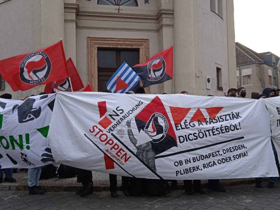 Bei einer Demonstration wird ein Banner gehalten; auf dem steht "NS-Verherrlichung stoppen". Dahinter sind viele Antifa-Fahnen und eine Fahne der VVN-BdA zu sehen.