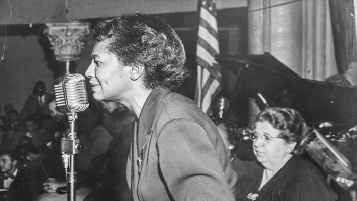 Claudia Jones spricht vor einem Publikum in ein Mikrofon. Im Hintergrund die amerikanische Flagge.