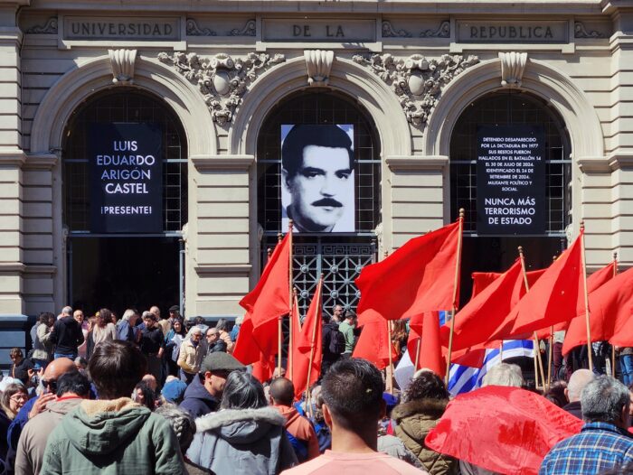 Menschenmenge mit roten Fahnen vor einem Gebäude der Universität in Montevideo. Über der Tür hängt die Aufschrift "Luis Eduardo Arigón Castel Presente", daneben ist ein Schwarzweißporträt von ihm zu sehen.